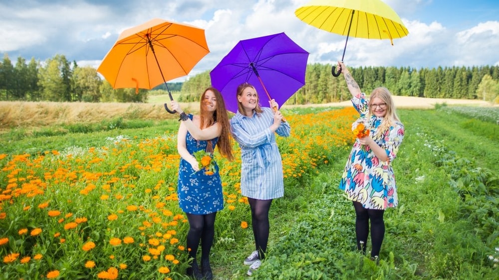 Kolme hymyilevää henkilöä seisoo kukkapellon keskellä ja pitelee käsissään värikkäitä sateenvarjoja.
