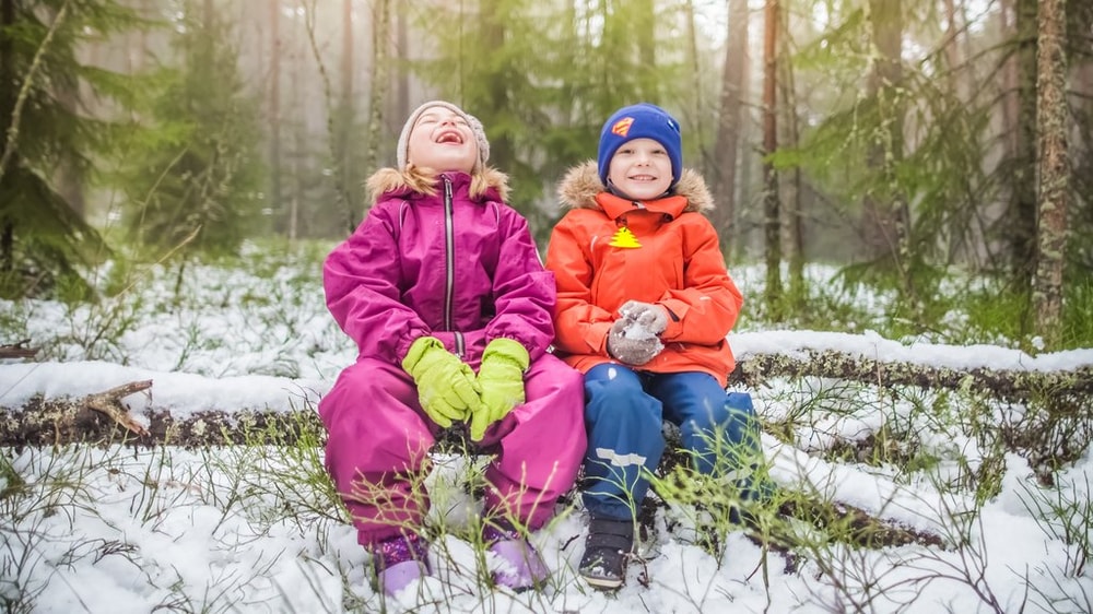 Kaksi lasta istuu lumisessa metsässä puunrungon päällä nauraen.
