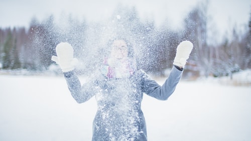 Tyttö seisoo lumisateessa ja heittää lunta päälleen nauraen