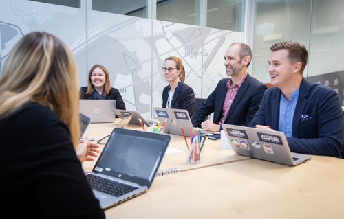 Ihmisiä pöydän ympärillä tietokoneiden ääressä, keskustelevat keskenään hymyillen